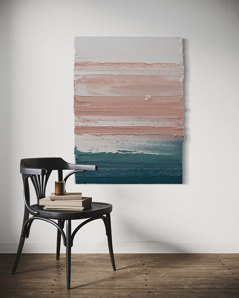 A peach, coral, white and teal with lavender thick textured painting by Teodora Guererra hangs on a white wall in a kitchen with a dark oak chair in front. Sitting on the chair are books and a candle.