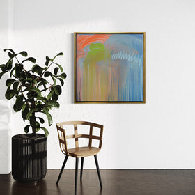 A blue, orange and green abstract print framed in a gold floater frame by Teodora Guererra hanging on a white wall with a large potted plant and chair in front.