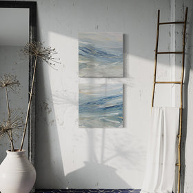 A pair of abstract coastal paintings with blue, grey, white, cream, stone, teal, and yellow ochre paint hanging on a white stucco wall by Teodora Guererra. To the right is a natural wood decorative ladder with a white fabric draped over it. to the left is a wall mirror with a dark ridged frame leaning on the wall and a large white ceramic pot with decorative sticks in front. The floor below is a dark blue tile in a geometric pattern with grey grout.