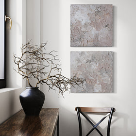 A pair of thickly textured abstract paintings in light pink, grey, coral and white by Teodora Guererra hanging on a white wall over a chair. To the left is a wood bench with a black ceramic vase holding tree branches in it. Beyond is a window.