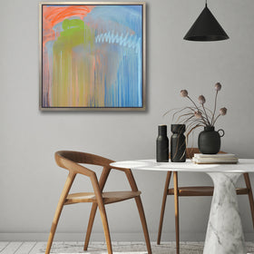 A blue, orange and green abstract print in a silver floater frame hangs on a white wall above two wood chairs and a round white marble pedestal table with black vases and a stack of books.