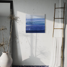 A blue and white textured abstract painting by Teodora Guererra hangs on a white wall. To the right is a natural wood decorative ladder with a white fabric draped over it. to the left is a wall mirror with a dark ridged frame leaning on the wall and a large white ceramic pot with decorative sticks in front. The floor below is a dark blue tile in a geometric pattern with grey grout.
