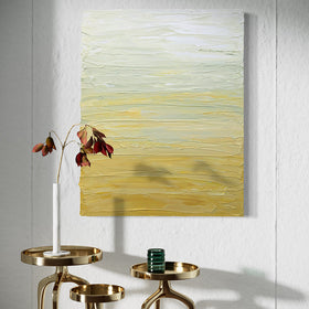 A Yellow, seafoam green and white textured painting by Teo Guererra hangs on a wall over 3 brass decorative tables with a sprig of orange leaves in a white vase and a green glass candle.
