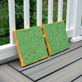 Two square abstract text based paintings with thick green paint and tangerine gestural lines, leaning against a white railing on a gray composite deck.