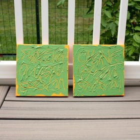 Two square abstract text based paintings with thick green paint and tangerine gestural lines, leaning against a white railing on a gray wooden deck with green foliage in the background.