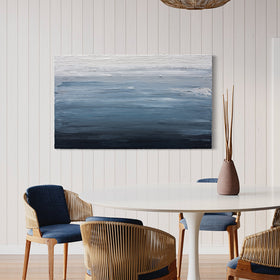 A thickly painted painting in dark navy blue, light blue and white by Teodora Guererra hang on a white wall in a kitchen. A white pedestal table surrounded by modern wicker type chairs with blue cushions sits in front of the paintings with a ceramic vase and sticks sitting on the table. Over the table hangs a wicker light and a door to the right can be seen in apple green.