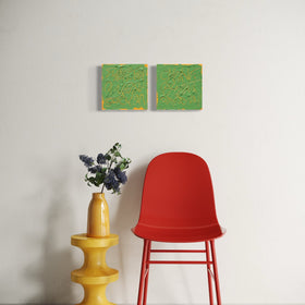 A modern red molded plastic chair with red metal legs is positioned next to a yellow tiered side table holding a yellow vase with purple flowers, against a white wall with two square abstract text based paintings with thick green paint and tangerine gestural lines, hanging above.