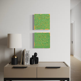 Two square abstract text based paintings with thick green paint and tangerine gestural lines, hanging on a white wall above a beige console table with a lamp and books.