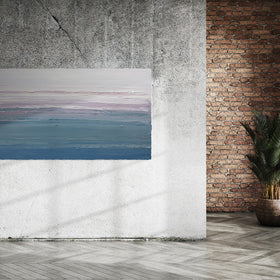 A white, lavender, teal and blue abstract heavily textured painting like sculpture by Teodora Guererra hangs on a grey cement wall with sunlight streaming  through the window in an industrial room.
