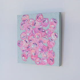 A very textured painting with celadon background and circle type abstract flowers in pink, peach, plum and white hangs on a white wall, side view.