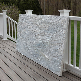 A white and light blue abstract painting with thick, impasto brushstrokes sits on a deck at an angle at the artists studio.