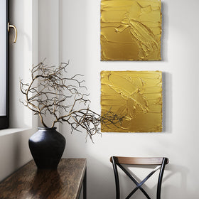 A pair of golden textured paintings by Teodora Guererra hanging on a wall above a black and brown chair. To the left is a wood table under a window with a black ceramic vase holding dried branches.