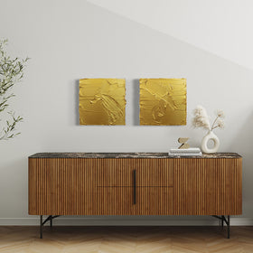 A pair of golden textured paintings by Teodora Guererra hang on a wall side by side above a mid century modern credenza in brown paneled wood with marble top. Sitting on the credenza is a white ceramic vase with dried white flowers and to the left are books with a small good sculpture. To the left of the credenza is a large potted fica tree and to the right of the credenza there are stairs leading up.