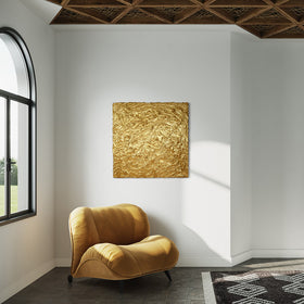 A metallic gold thick textured painting on a white wall in natural light by Teodora Guererra hangs over a yellow ochre velvet modern chair. To the left and right are windows and in front is a carpet in a black and white pattern.
