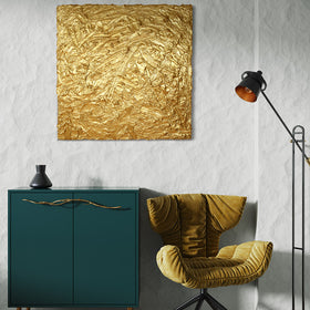 A metallic gold thick textured painting on a white wall in natural light by Teodora Guererra hangs over a green cabinet with brass tree branch like handles and a black ceramic vase sits on top. To the right is a yellow ochre velvet modern chair and a black floor lamp.