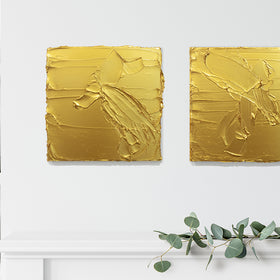 A pair of golden textured paintings by Teodora Guererra hang on a wall above a white shelf. To the left are more shelves holding a basket, small plant, coral and other items. Under the paintings on the shelf is a white ceramic vase with a green plant.