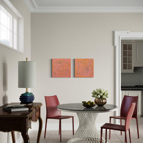 A dining area featuring a round gray-top table with a white woven pedestal base, surrounded by two reddish-brown chairs. The room has beige walls, two abstract orange and pink paintings, a dark wood console table with a lamp, and a doorway leading to a kitchen.