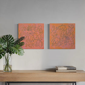 Two square abstract text based paintings with orange-pink backgrounds and thin teal scribbled lines, displayed side-by-side on a light gray wall above a wooden console table with a green plant and books. A large arched window on the right shows a city skyline and patio chair.