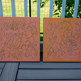 Two square abstract text based paintings with orange and peach backgrounds and teal scribbled line art, displayed side-by-side on a dark gray wooden deck in front of a white railing with green foliage in the background.