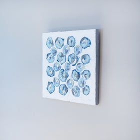 A very textured abstract painting with white background and circle type abstract flowers in blue, teal, green, plum and white hangs on a grey wall.