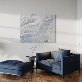 A blue, grey, white, stone, teal, and yellow ochre thickly textured abstract painting by Teodora Guererra on a white wall in natural light hangs over a blue oversized chair with ottoman. A contemporary end table is to the side of the chair with a modern light. Also, a plant in a grey pot sits on the floor to the right of the blue chair.