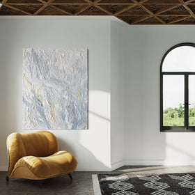 An abstract heavily textured painting with blue, grey, white, stone, teal, and yellow ochre hangs on a white wall in natural light by Teodora Guererra. This painting hangs over a yellow ochre velvet modern chair. To the left and right are windows and in front is a carpet in a black and white pattern. To the extreme right is a plant of lavender in a black ribbed pot.