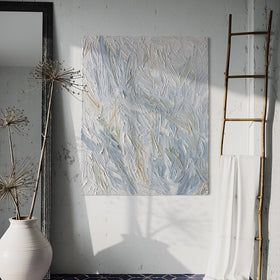 An abstract heavily textured painting with blue, grey, white, stone, teal, and yellow ochre paint hanging on a white stucco wall by Teodora Guererra. To the right is a natural wood decorative ladder with a white fabric draped over it. to the left is a wall mirror with a dark ridged frame leaning on the wall and a large white ceramic pot with decorative sticks in front. The floor below is a dark blue tile in a geometric pattern with grey grout.
