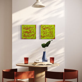 Two square abstract text-based paintings with textured chartreuse over hot raspberry pink  graffiti-style lettering, hang on a light gray wall above a round dining table with two rust-red chairs. A blue vase with a green leaf and an open book sit on the table, with sunlight casting shadows across the wall.