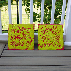 Two square abstract text-based paintings with textured chartreuse over hot raspberry pink  graffiti-style lettering, displayed side-by-side on a gray wooden deck in front of a white railing with green foliage in the background.