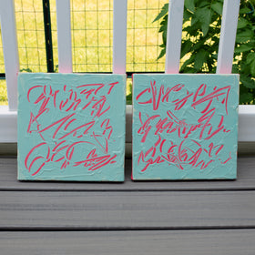 Two square texted based abstract paintings with textured teal backgrounds and pink abstract graffiti-style lettering, displayed side-by-side on a gray wooden deck in front of a white fence with green foliage in the background.