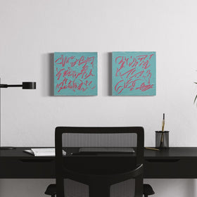 Two square abstract text based paintings with pink carved writing on a teal background, displayed side-by-side on a white wall above a black desk setup with a chair, lamp, and plant.