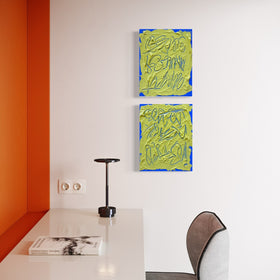 Two abstract text based paintings feature a thick, yellow-green impasto texture with blue abstract scribbled lines and a bright blue base and hang on a white wall in a modern room with an orange accent wall, white desk, and gray chair.