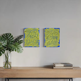 Two abstract text based painting featuring a thick, yellow-green impasto texture with blue abstract scribbled lines hang on a light gray wall above a wooden console table with a potted plant and books. A large arched window on the right shows a city view and outdoor chair.