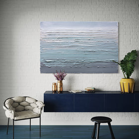 A white, teal and grey heavily textured abstract painting by Teodora Guererra hangs on a white brick wall in natural light over a blue credenza with some vases of flowers, books and a yellow pot with a green plant. To the left is a contemporary chair in a stone fabric and a black stool in front.