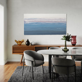 A blue, light pink, coral and white thickly textured abstract painting hanging on a white wall by Teodora Guererra hangs in a dining room over a mid century cabinet with assorted vases and stoneware. The dining chairs a grey modern fabric with black iron legs and the table of white marble top with black legs sits on a black and white nubby carpet on an oak floor.