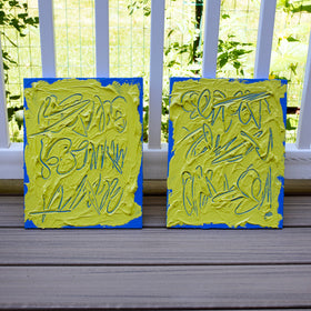 Two abstract text based painting featuring a thick, yellow-green impasto texture with blue abstract scribbled lines and a bright blue background. They are displayed on a wooden deck in front of a white railing with green foliage behind.