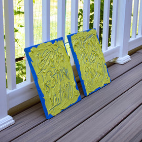 Two abstract text based painting featuring a thick, yellow-green impasto texture with blue abstract scribbled lines and a bright blue background. They are displayed on a wooden deck, side view, in front of a white railing with green foliage behind.