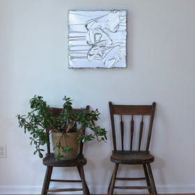 A silver metallic textured painting by Teodora Guererra hanging on a wall in the artists studio above 2 wooden chairs, one with a yucca plant.