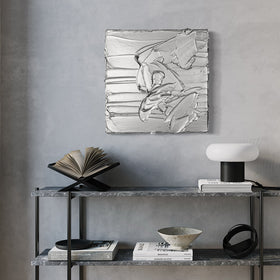 A silver metallic textured painting by Teodora Guererra hanging on a wall above a marble shelf unit with books and decorative objects.
