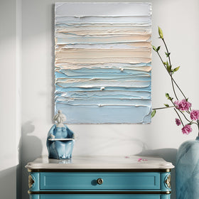 A thickly painted painting in white, orange sherbet and teal blue by Teodora Guererra hangs on a white wall over an ornate teal end table with white marble top. To the right is a marble vase with pink flowers and sitting on the table is a figurine of an asian woman in a teal dress. The painting is like a wall sculpture.