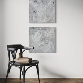 A pair of thickly textured paintings in grey, white, blue grey and plum grey by Teodora Guererra hanging on a white wall in front of a brown chair with books and a candle sitting on it.