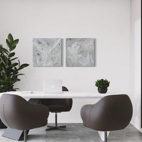 A pair of thickly textured paintings in grey, white, blue grey and plum grey by Teodora Guererra hanging on a white wall behind a desk.