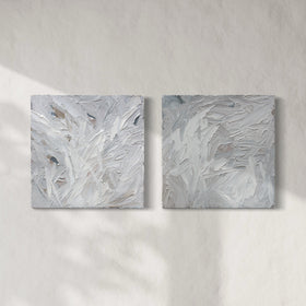 A pair of thickly textured paintings in grey, white, blue grey and plum grey by Teodora Guererra hanging on a white wall.