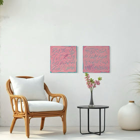 Two square pink text based paintings with writing into the surface with various tools, carving fluid script that reveals the teal color hidden below. Displayed side-by-side on a white wall in a room with wicker chair and table with flowers.