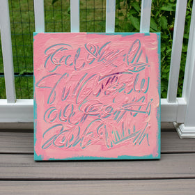 A square painting with a pink background and textured abstract lettering, leaning against a white railing on a wooden deck with green foliage in the background.