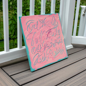 A square canvas wall art piece leaning on a gray composite deck next to a white railing, featuring a pink background with textured turquoise paint and cursive script lettering.