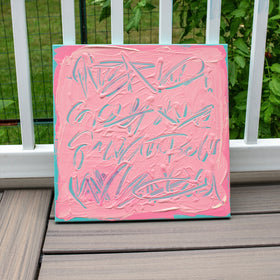 A square canvas painting with thick pink paint and abstract light blue and white brushstrokes, leaning against a white railing on a wooden deck with green foliage in the background.