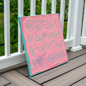 A square canvas art print with thick pink and teal paint textures and abstract lettering, leaning against a white wooden deck railing with green foliage in the background.