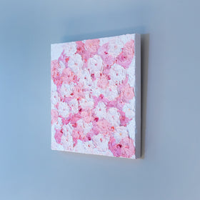 A very textured abstract painting with white background and circle type abstract flowers in pink and white hangs on a grey wall, side view.