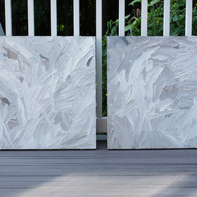 A pair of thickly textured paintings in grey, white, blue grey and plum grey by Teodora Guererra leaning outside on a deck.